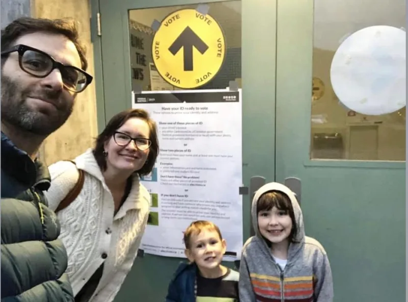 Two adults and two young children smile for a selfie at the door of a polling station, beside a yellow Vote sign with an arrow.