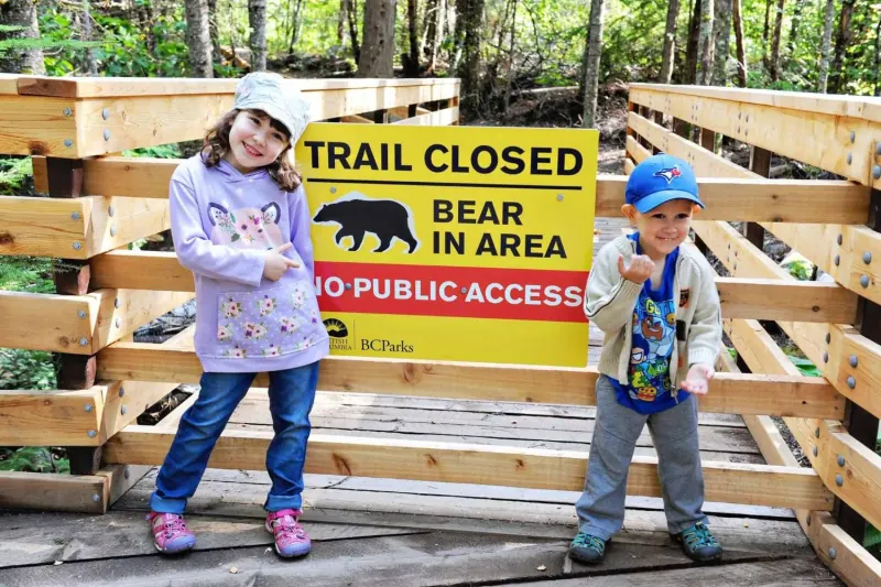 Two young children pose beside a yellow BC Parks sign on a wooden boardwalk in the forest. The sign reads Trail closed, bear in area, no public access.